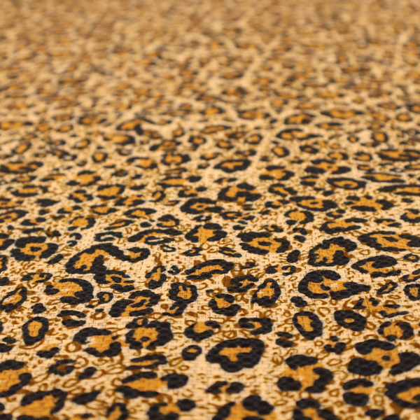 The design consists of irregular, organic black "rosette" shapes with golden-tan centres, set against a warm, sandy-beige background, creating a style of leopard print. The fabric has a visible textured, canvas-like weave, giving the pattern a slightly rugged and tactile appearance.