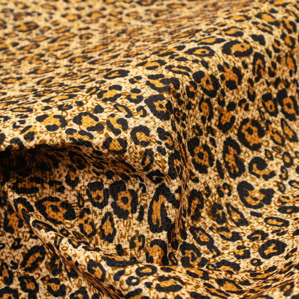 The design consists of irregular, organic black "rosette" shapes with golden-tan centres, set against a warm, sandy-beige background, creating a style of leopard print. The fabric has a visible textured, canvas-like weave, giving the pattern a slightly rugged and tactile appearance.
