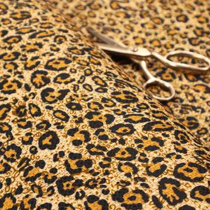 The design consists of irregular, organic black "rosette" shapes with golden-tan centres, set against a warm, sandy-beige background, creating a style of leopard print. The fabric has a visible textured, canvas-like weave, giving the pattern a slightly rugged and tactile appearance.