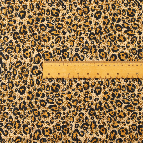 The design consists of irregular, organic black "rosette" shapes with golden-tan centres, set against a warm, sandy-beige background, creating a style of leopard print. The fabric has a visible textured, canvas-like weave, giving the pattern a slightly rugged and tactile appearance.