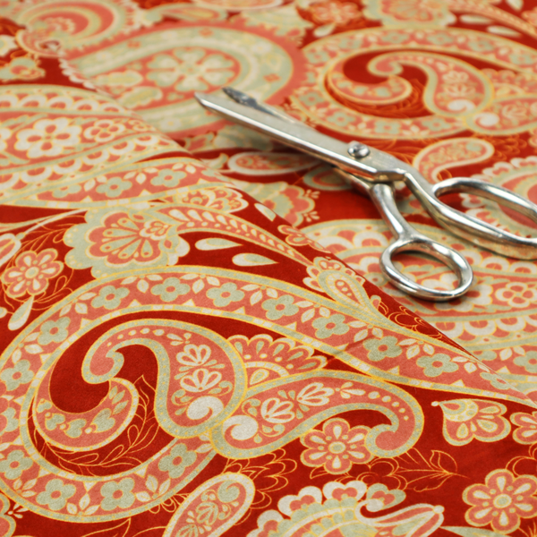 A vibrant, warm-toned textile featuring large-scale paisley teardrop motifs in shades of cream, pale mint, and soft orange against a bold red background. The intricate patterns within the paisley shapes include delicate floral borders and internal mandalas, surrounded by smaller scattered flower accents for a rich, traditional Indian-inspired aesthetic.