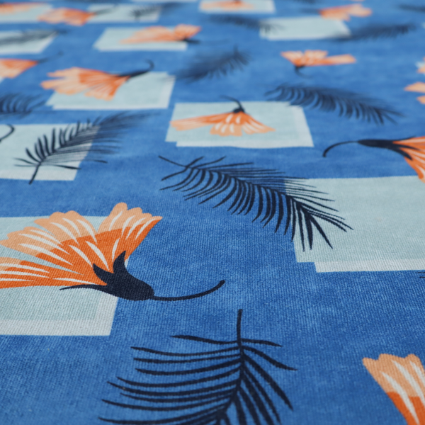 Modern tropical fabric featuring a vibrant cobalt blue background with scattered orange fan-shaped flowers, dark navy palm fronds, and pale blue geometric square overlays.