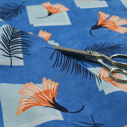 Modern tropical fabric featuring a vibrant cobalt blue background with scattered orange fan-shaped flowers, dark navy palm fronds, and pale blue geometric square overlays.