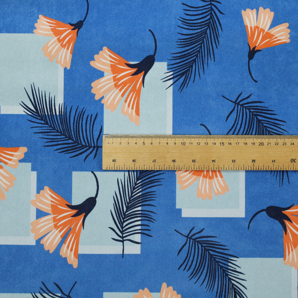 Modern tropical fabric featuring a vibrant cobalt blue background with scattered orange fan-shaped flowers, dark navy palm fronds, and pale blue geometric square overlays.
