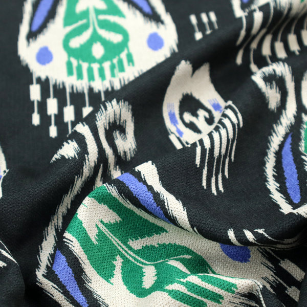 A dark textile featuring rows of floating, feathered ikat medallions. The design glows with emerald green and bright sapphire blue against a deep obsidian background, accented by crisp white geometric tassels and swirling crown-like scrollwork.