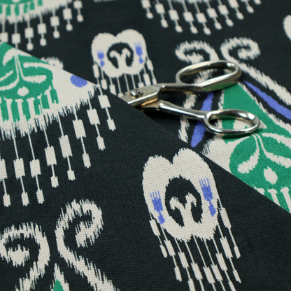 A dark textile featuring rows of floating, feathered ikat medallions. The design glows with emerald green and bright sapphire blue against a deep obsidian background, accented by crisp white geometric tassels and swirling crown-like scrollwork.