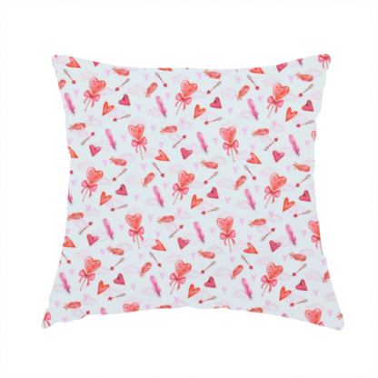 The fabric used to create this handmade cushion is a white fabric featuring a light, whimsical Valentine’s Day pattern with watercolour elements scattered across a pale background. The design includes pink heart-shaped balloons tied with bows, soft pink feathers, and various small arrows and hearts. Faint, dreamy cloud-like textures in pink and blue are visible in the background, adding to the soft, airy aesthetic.