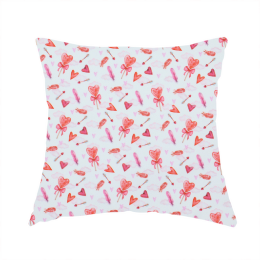 The fabric used to create this handmade cushion is a white fabric featuring a light, whimsical Valentine’s Day pattern with watercolour elements scattered across a pale background. The design includes pink heart-shaped balloons tied with bows, soft pink feathers, and various small arrows and hearts. Faint, dreamy cloud-like textures in pink and blue are visible in the background, adding to the soft, airy aesthetic.