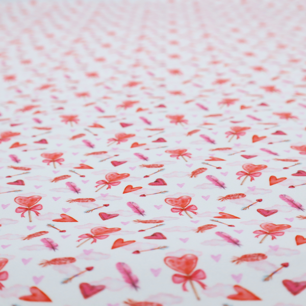 This white fabric features a light, whimsical Valentine’s Day pattern with watercolour elements scattered across a pale background. The design includes pink heart-shaped balloons tied with bows, soft pink feathers, and various small arrows and hearts. Faint, dreamy cloud-like textures in pink and blue are visible in the background, adding to the soft, airy aesthetic.