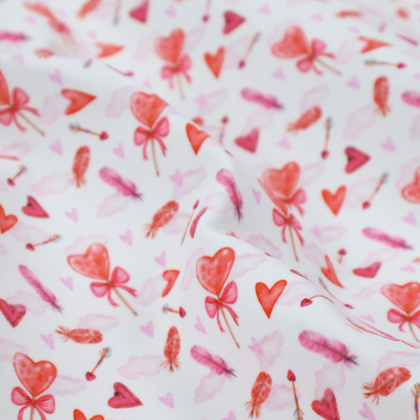 This white fabric features a light, whimsical Valentine’s Day pattern with watercolour elements scattered across a pale background. The design includes pink heart-shaped balloons tied with bows, soft pink feathers, and various small arrows and hearts. Faint, dreamy cloud-like textures in pink and blue are visible in the background, adding to the soft, airy aesthetic.