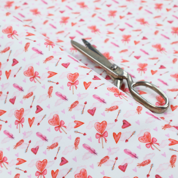 This white fabric features a light, whimsical Valentine’s Day pattern with watercolour elements scattered across a pale background. The design includes pink heart-shaped balloons tied with bows, soft pink feathers, and various small arrows and hearts. Faint, dreamy cloud-like textures in pink and blue are visible in the background, adding to the soft, airy aesthetic.