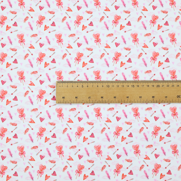 This white fabric features a light, whimsical Valentine’s Day pattern with watercolour elements scattered across a pale background. The design includes pink heart-shaped balloons tied with bows, soft pink feathers, and various small arrows and hearts. Faint, dreamy cloud-like textures in pink and blue are visible in the background, adding to the soft, airy aesthetic.