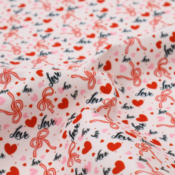 This light pink fabric features a dense, repeating Valentine’s Day pattern with a vintage, romantic aesthetic. The design is composed of small red hearts and larger pink hearts, interspersed with delicate red ribbon bows and the word "love" written in a dark, flowing cursive script. Tiny red dots are scattered throughout the background, creating a textured, confetti-like effect across the entire print.