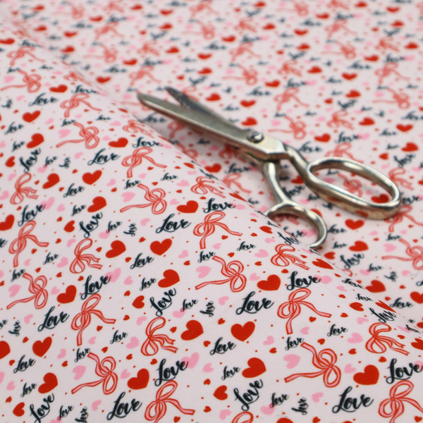 This light pink fabric features a dense, repeating Valentine’s Day pattern with a vintage, romantic aesthetic. The design is composed of small red hearts and larger pink hearts, interspersed with delicate red ribbon bows and the word "love" written in a dark, flowing cursive script. Tiny red dots are scattered throughout the background, creating a textured, confetti-like effect across the entire print.