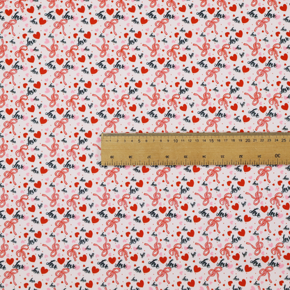 This light pink fabric features a dense, repeating Valentine’s Day pattern with a vintage, romantic aesthetic. The design is composed of small red hearts and larger pink hearts, interspersed with delicate red ribbon bows and the word "love" written in a dark, flowing cursive script. Tiny red dots are scattered throughout the background, creating a textured, confetti-like effect across the entire print.