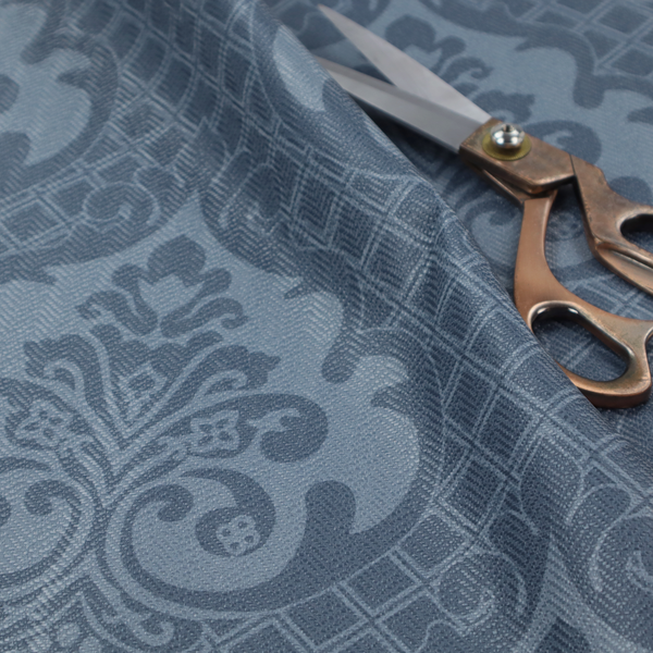 A dusty blue and steel grey damask pattern on a textured fabric. Large, ornate floral motifs with stylized acanthus leaves are arranged in a repeating grid. These medallions are a lighter, cool-toned blue-grey set against a darker navy-blue background.