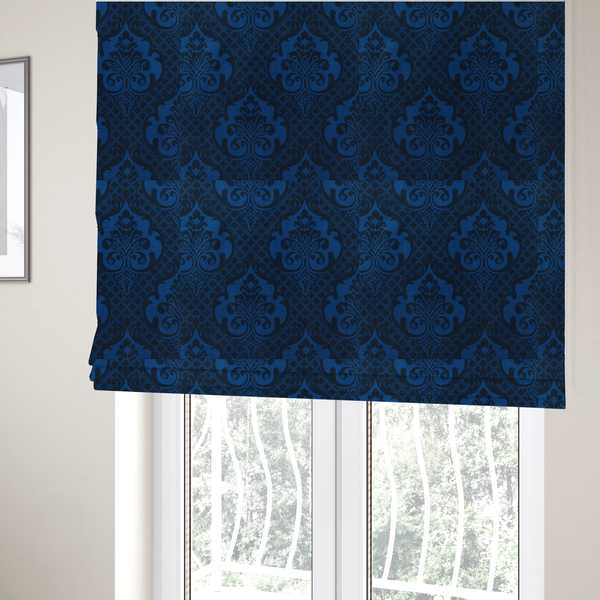 The fabric used to create these Roman blinds has a design that features large, ornate floral medallions and scrolling acanthus leaves in a bright royal blue shade, set against a deep navy background. A fine diagonal lattice grid fills the space between the motifs, and the material displays a clear woven texture that adds structural depth and a subtle sheen to the tonal blue design.