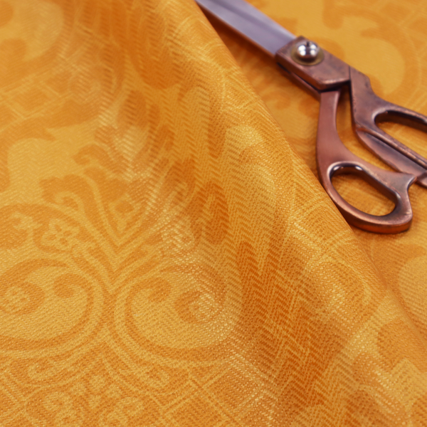 An orange damask pattern on a textured fabric. Large, ornate floral motifs and scrolling acanthus leaves are arranged in a repeating grid. These motifs are rendered in a soft peach shade against a more saturated, vibrant orange background.