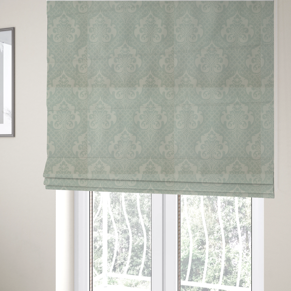 The fabric used to create these Roman blinds has a design that showcases large, ornate floral medallions and scrolling acanthus leaves in a light silver shade, set against a slightly darker grey-white background. A fine diagonal lattice grid creates a diamond pattern in the negative space between the motifs, and the material displays a clear woven texture that adds a sophisticated, metallic-like shimmer to the pattern.