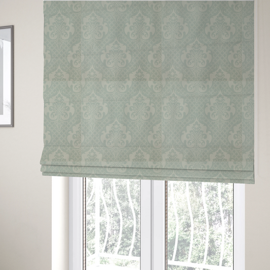 The fabric used to create these Roman blinds has a design that showcases large, ornate floral medallions and scrolling acanthus leaves in a light silver shade, set against a slightly darker grey-white background. A fine diagonal lattice grid creates a diamond pattern in the negative space between the motifs, and the material displays a clear woven texture that adds a sophisticated, metallic-like shimmer to the pattern.