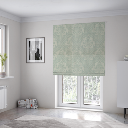 The fabric used to create these Roman blinds has a design that showcases large, ornate floral medallions and scrolling acanthus leaves in a light silver shade, set against a slightly darker grey-white background. A fine diagonal lattice grid creates a diamond pattern in the negative space between the motifs, and the material displays a clear woven texture that adds a sophisticated, metallic-like shimmer to the pattern.