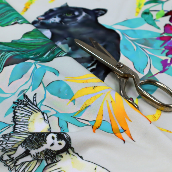 A vibrant tropical fabric print featuring sleek black panthers prowling among exotic flora. The design includes colourful botanical elements like oversized teal and lime green leaves, magenta flowers, and yellow ferns. Small details like a soaring owl and a hummingbird are scattered throughout on a pale grey background.
