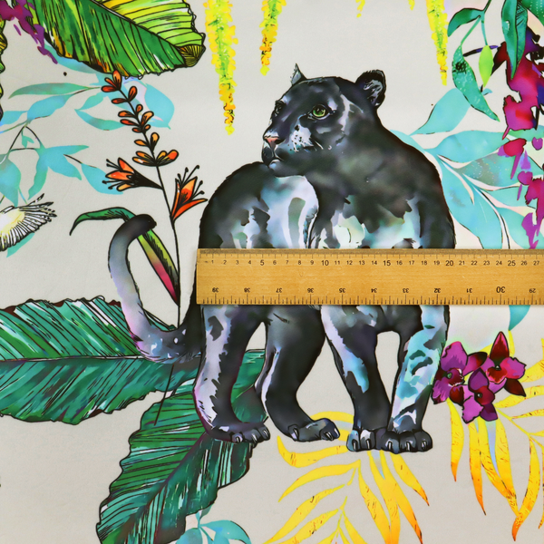A vibrant tropical fabric print featuring sleek black panthers prowling among exotic flora. The design includes colourful botanical elements like oversized teal and lime green leaves, magenta flowers, and yellow ferns. Small details like a soaring owl and a hummingbird are scattered throughout on a pale grey background.