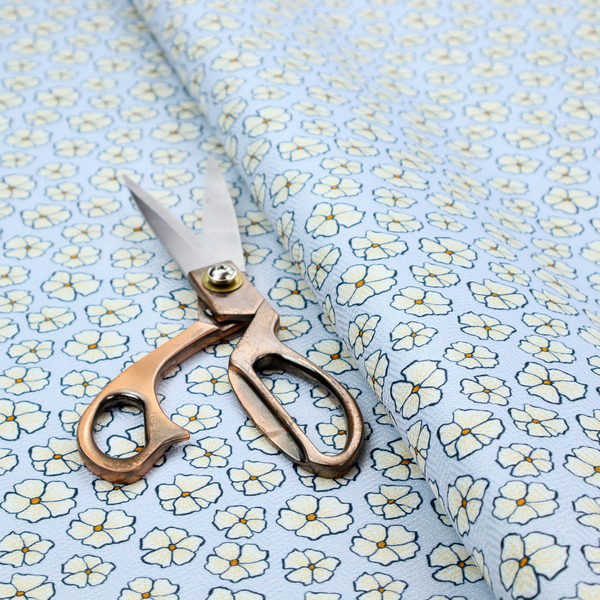 A light blue fabric featuring a dense, repeating pattern of small white flowers with yellow centres and dark outlines.