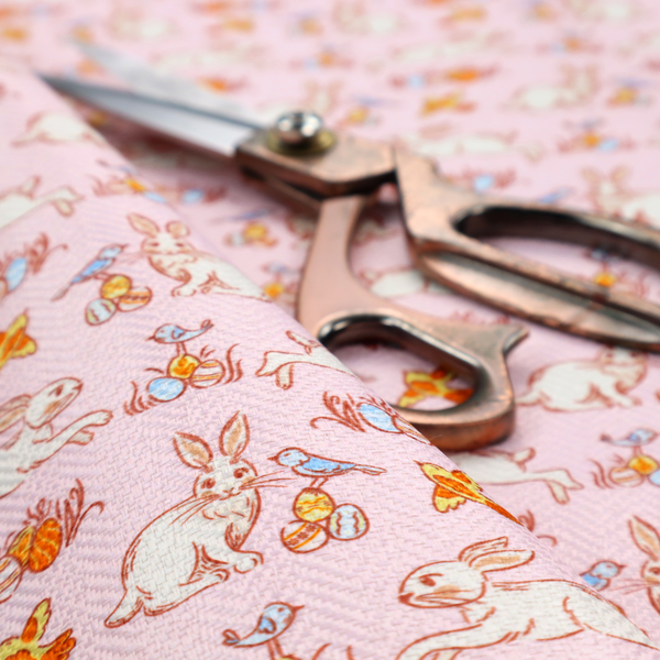 This fabric features a vintage-style Easter meadow pattern on a soft, blush-pink background. The design is reminiscent of classic children’s book illustrations, using fine, orange-brown linework and a muted pastel palette.