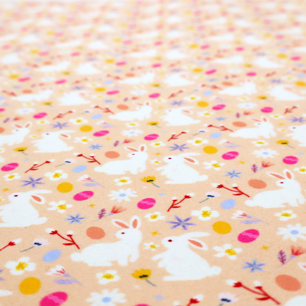 This fabric features a delicate, dainty Easter meadow pattern on a soft, peachy-beige background. The design is airy and feminine, characterized by small-scale illustrations and a palette of pastel and vibrant spring colours.