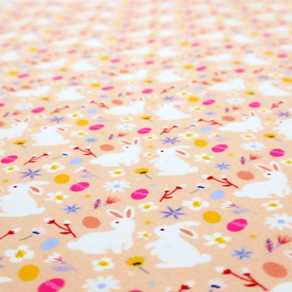 This fabric features a delicate, dainty Easter meadow pattern on a soft, peachy-beige background. The design is airy and feminine, characterized by small-scale illustrations and a palette of pastel and vibrant spring colours.