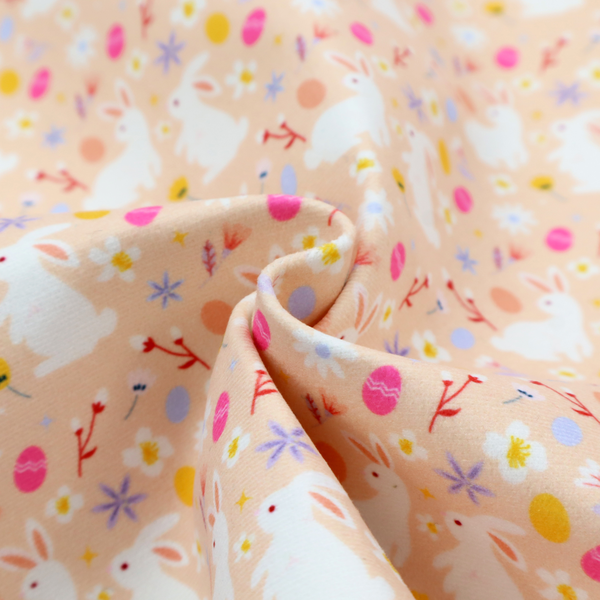 This fabric features a delicate, dainty Easter meadow pattern on a soft, peachy-beige background. The design is airy and feminine, characterized by small-scale illustrations and a palette of pastel and vibrant spring colours.