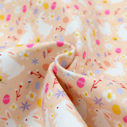 This fabric features a delicate, dainty Easter meadow pattern on a soft, peachy-beige background. The design is airy and feminine, characterized by small-scale illustrations and a palette of pastel and vibrant spring colours.