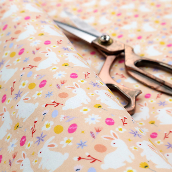 This fabric features a delicate, dainty Easter meadow pattern on a soft, peachy-beige background. The design is airy and feminine, characterized by small-scale illustrations and a palette of pastel and vibrant spring colours.