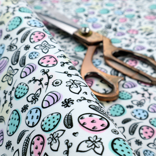 This fabric features an artistic, hand-illustrated Easter egg pattern on a cool, pale blue background. The design has a sketchbook or "doodle" quality, with expressive black ink outlines and soft watercolour-style colour accents.