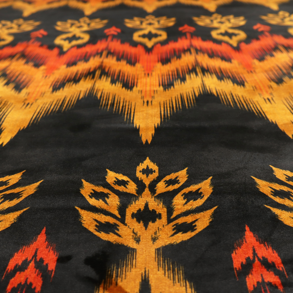 A bold ethnic-inspired fabric featuring a horizontal Ikat border design. The pattern showcases jagged, flame-like chevron stripes in vibrant orange and saffron yellow, flanked by rows of stylized tribal leaf motifs on a dark charcoal-grey background.