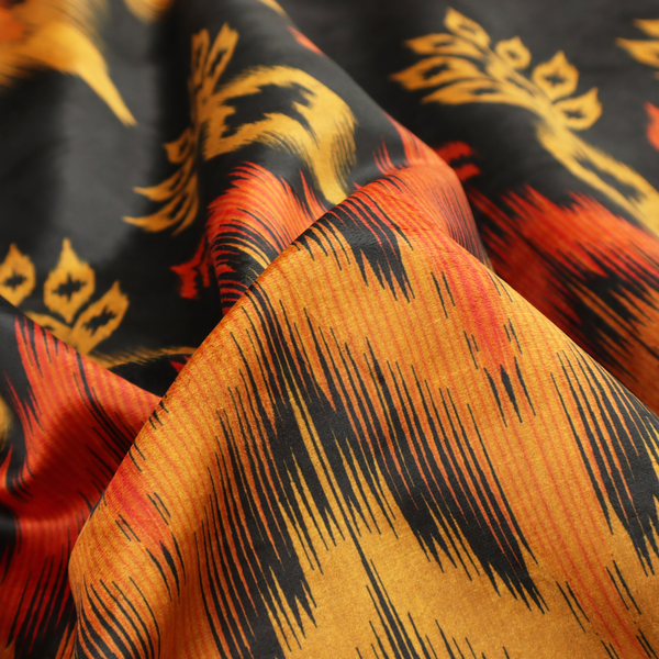 A bold ethnic-inspired fabric featuring a horizontal Ikat border design. The pattern showcases jagged, flame-like chevron stripes in vibrant orange and saffron yellow, flanked by rows of stylized tribal leaf motifs on a dark charcoal-grey background.