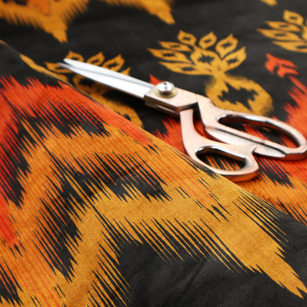 A bold ethnic-inspired fabric featuring a horizontal Ikat border design. The pattern showcases jagged, flame-like chevron stripes in vibrant orange and saffron yellow, flanked by rows of stylized tribal leaf motifs on a dark charcoal-grey background.