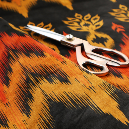 A bold ethnic-inspired fabric featuring a horizontal Ikat border design. The pattern showcases jagged, flame-like chevron stripes in vibrant orange and saffron yellow, flanked by rows of stylized tribal leaf motifs on a dark charcoal-grey background.