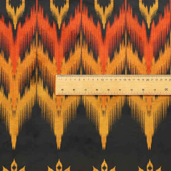 A bold ethnic-inspired fabric featuring a horizontal Ikat border design. The pattern showcases jagged, flame-like chevron stripes in vibrant orange and saffron yellow, flanked by rows of stylized tribal leaf motifs on a dark charcoal-grey background.