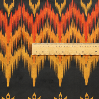 A bold ethnic-inspired fabric featuring a horizontal Ikat border design. The pattern showcases jagged, flame-like chevron stripes in vibrant orange and saffron yellow, flanked by rows of stylized tribal leaf motifs on a dark charcoal-grey background.