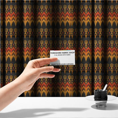 The fabric used to create these made-to-measure curtains is a bold ethnic-inspired fabric featuring a horizontal Ikat border design. The pattern showcases jagged, flame-like chevron stripes in vibrant orange and saffron yellow, flanked by rows of stylized tribal leaf motifs on a dark charcoal-grey background.