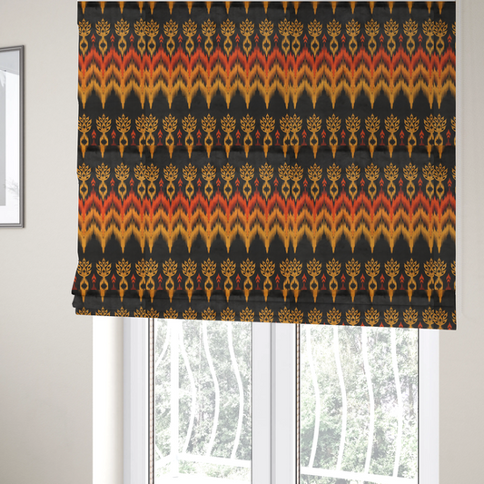 The fabric used to create these Roman blinds is a bold ethnic-inspired fabric featuring a horizontal Ikat border design. The pattern showcases jagged, flame-like chevron stripes in vibrant orange and saffron yellow, flanked by rows of stylized tribal leaf motifs on a dark charcoal-grey background.