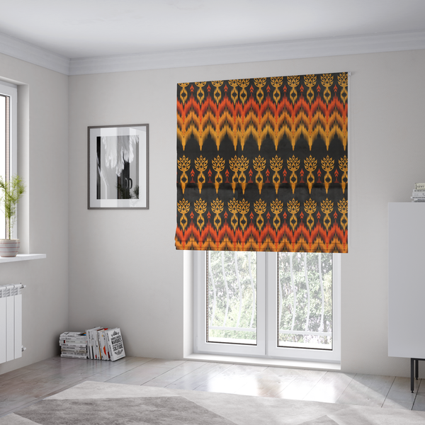 The fabric used to create these Roman blinds is a bold ethnic-inspired fabric featuring a horizontal Ikat border design. The pattern showcases jagged, flame-like chevron stripes in vibrant orange and saffron yellow, flanked by rows of stylized tribal leaf motifs on a dark charcoal-grey background.