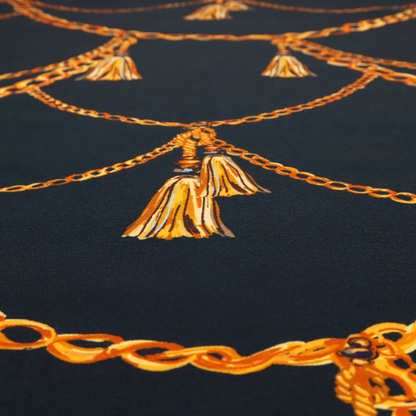 A luxury fashion-inspired fabric on a deep navy-black background. The design features elegant, draped golden chains arranged in a repeating scallop or 'swag' pattern, accented by highly detailed golden silk tassels suspended from ornate beaded cords.