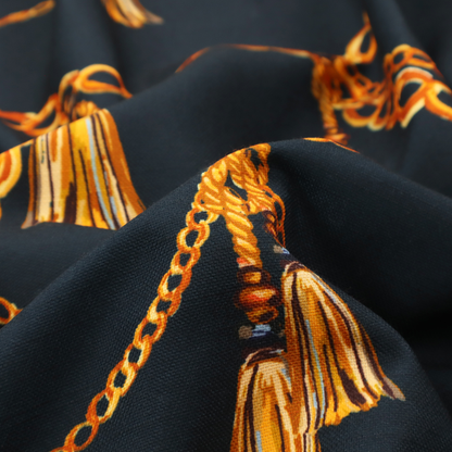 A luxury fashion-inspired fabric on a deep navy-black background. The design features elegant, draped golden chains arranged in a repeating scallop or 'swag' pattern, accented by highly detailed golden silk tassels suspended from ornate beaded cords.