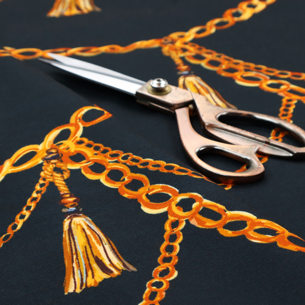 A luxury fashion-inspired fabric on a deep navy-black background. The design features elegant, draped golden chains arranged in a repeating scallop or 'swag' pattern, accented by highly detailed golden silk tassels suspended from ornate beaded cords.