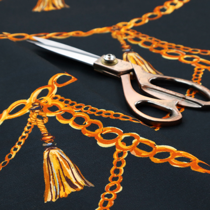 A luxury fashion-inspired fabric on a deep navy-black background. The design features elegant, draped golden chains arranged in a repeating scallop or 'swag' pattern, accented by highly detailed golden silk tassels suspended from ornate beaded cords.