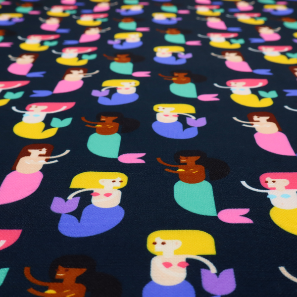 A fun and vibrant children's fabric on a deep midnight-navy background. The pattern features a diverse cast of illustrated mermaids with various skin tones, hair colours (pink, blonde, brown), and colourful tails in shades of mint, yellow, pink, and purple, arranged in a cheerful repeating grid.