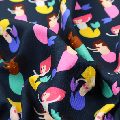 A fun and vibrant children's fabric on a deep midnight-navy background. The pattern features a diverse cast of illustrated mermaids with various skin tones, hair colours (pink, blonde, brown), and colourful tails in shades of mint, yellow, pink, and purple, arranged in a cheerful repeating grid.